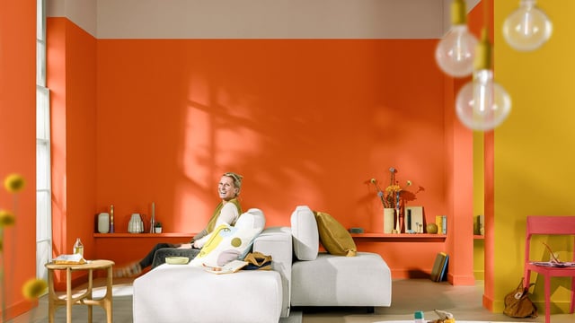 Moodne elutuba oranžide seintega; inimene lõõgastub voodil, värvikas sisustus ja taimed.